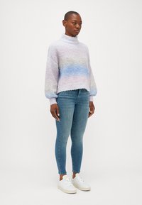 Pastelna skrajšana pulover z visokim ovratnikom in balonskimi rokavi, ki ima gradient od sivke do modre, v kombinaciji s prilegajočimi modrimi kavbojkami in belimi supergami.