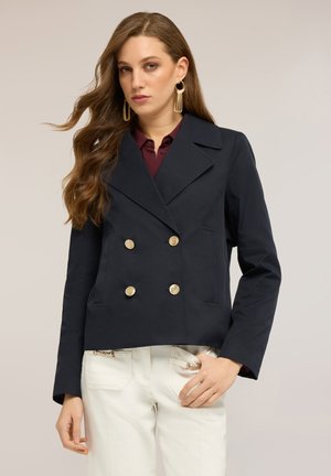 Femme aux longs cheveux portant des boucles d'oreilles à pampilles en forme d'anneaux dorés, un blazer croisé sombre avec des boutons dorés, et un pantalon blanc avec des détails zippés.