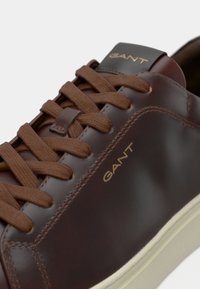 Sneaker din piele maro cu sireturi plate maro, logo-ul auriu "GANT" pe lateral și talpă din cauciuc crem. Textură netedă cu detalii de cusătură subtile.