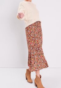 Světlý béžový pletený svetr s výrazným velkým vzorem pletení, doplněný dlouhou vzorovanou sukní v teplých tónech. Kompletní vzhled doplňují hnědé kotníkové boty.