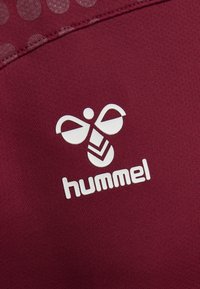 Burgundy sportskjorta med strukturerad tyg, med en vit logotyp av en stiliserad bi och ordet "hummel" i fet stil under.