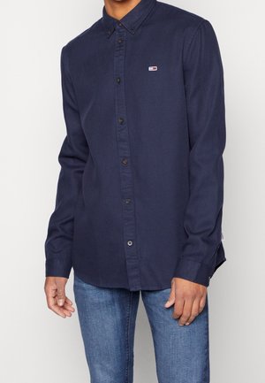 Muž v tmavě modré košili s dlouhým rukávem na knoflíky s malým logem na hrudi a modrých džínách, stojící na jednobarevném pozadí.