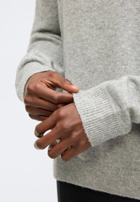 Χέρια που ρυθμίζουν το μανίκι ενός ανοιχτόχρωμου γκρι πουλόβερ από μαλλί, φορεμένο πάνω από μαύρο παντελόνι.