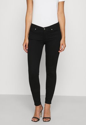 Kvinde iført sorte skinny jeans og sorte højhælede sandaler, kombineret med en hvid top med korte ærmer, stående mod en ensfarvet baggrund.