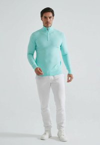 Pull léger turquoise avec un col zippé haut, design ajusté, poignets et ourlet côtelés, associé à un pantalon blanc slim et des baskets blanches.