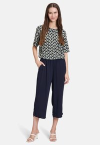 Wzorzysty top z krótkim rękawem w kolorze granatowym, zielonym i kremowym, o luźnym kroju; zestawiony z granatowymi spodniami o szerokich nogawkach; minimalistyczne sandały na cienkich obcasach.