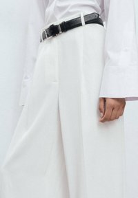 Pantalon blanc sur mesure avec une finition lisse, associé à une ceinture en cuir noire avec une boucle argentée. Des manches longues blanches dépassent de la chemise.