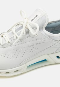 Hvit golfskoin med perforert design, teksturert skinnoverdel, lyseblå detaljer og en polstret geometrisk såle for bedre stabilitet.