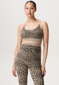 Vrouw met donker haar in een strakke knot draagt een beige en zwarte luipaardprint sportbeha en bijpassende high-waisted leggings, staande tegen een effen achtergrond.
