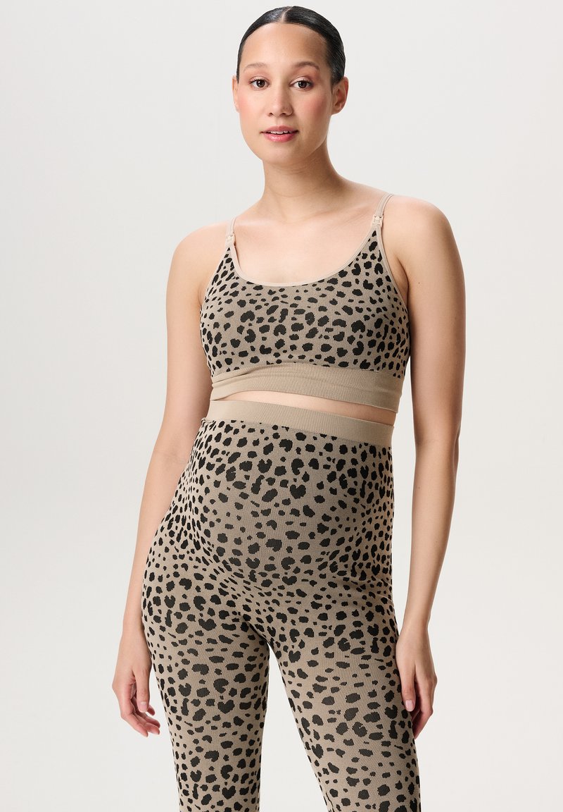Vrouw met donker haar in een strakke knot draagt een beige en zwarte luipaardprint sportbeha en bijpassende high-waisted leggings, staande tegen een effen achtergrond.