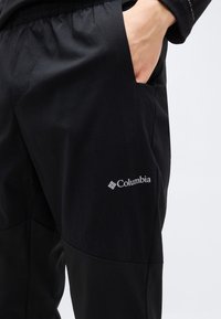 Χέρι στην τσέπη, φοράει μαύρο παντελόνι Columbia με ελαστική μέση και εμφανές λογότυπο Columbia στον μηρό, σε απλό φόντο.