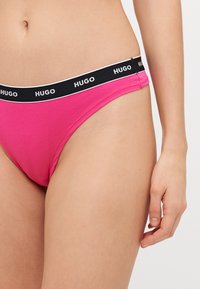 Culottes bikini en coton rose avec une ceinture logo noire ornée d'un passepoil blanc. La découpe sur les côtés offre un ajustement moderne et une couverture minimale.