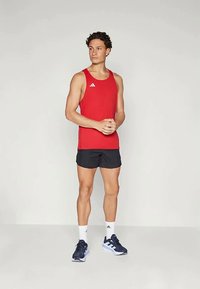 Piros sportos ujjatlan felső Adidas logóval, fekete rövidnadrág, fehér bokazokni és kék sportcipő. A modell egyenesen áll, kezét összekulcsolva.