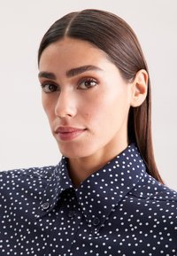 Camicia azzurra a pois con colletto abbottonato, caratterizzata da pois bianchi, una superficie liscia e bottoni blu sulla patta anteriore.