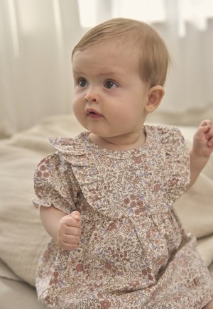 Baby with light hair wearing a floral print dress, sitting on a beige blanket, looking slightly to the side.