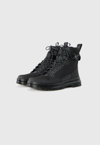 Botas negras de caña alta con paneles texturizados, cordones en el frente, correas con hebillas, suelas gruesas y tiradores, sobre un fondo blanco.