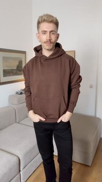 Brown oversized hoodie with a kangaroo pocket, features a subtle logo on the cuff. Paired with black pants. Minimalist background.