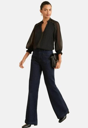Svart genomskinlig blus med silvernitar, i kombination med mörkblå utsvängda jeans. Modellen håller en svart handväska och har på sig spetsiga svarta skor.