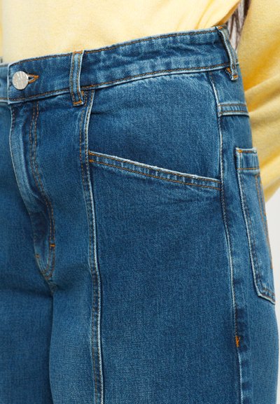 Blue high-waisted jeans with front and side pockets, orange stitching, and a silver button paired with a pale yellow top.