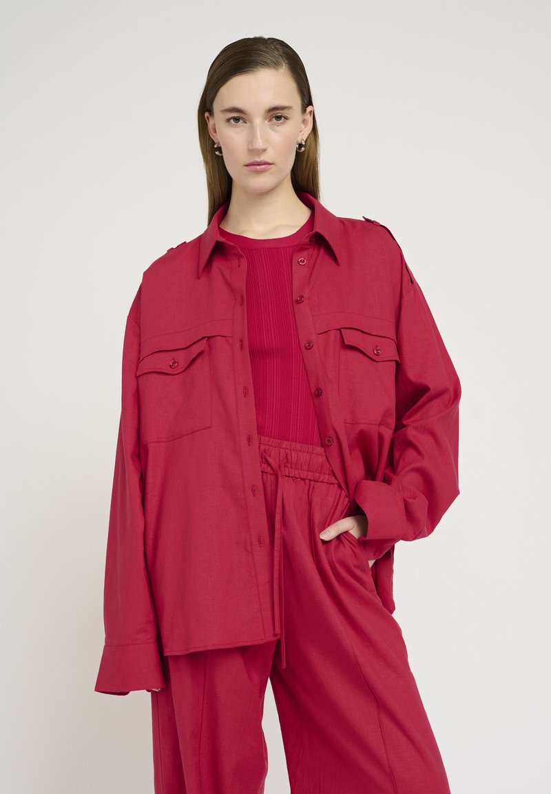 Camisa roja de botones con dos bolsillos en el pecho, combinada con una blusa roja de canalé y pantalones rojos holgados. Tejido suave, ajuste relajado.