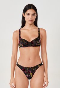 Donna che indossa un set di lingerie nera trasparente con ricami floreali rossi, rosa e gialli, in piedi contro uno sfondo chiaro e semplice.