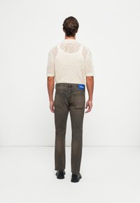 Egy beige hálós, rövid ujjú felsőt és elhalványult fekete farmert viselő személy, aki háttal áll egy fehér háttér előtt.