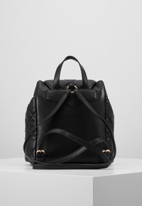 Mustat tilkkutäkkikuvioidut nahkareput säädettävillä hartioilla ja kultasävyisillä yksityiskohdilla valkoisella jalustalla harmaata taustaa vasten.