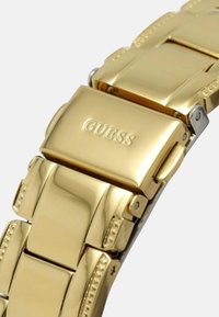 Zlatý nerezový hodinkový náramok s leštenými článkami, textúrovanými akcentmi a spona s vyrazeným názvom značky "GUESS."