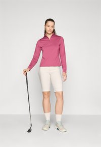 Maglia rosa a maniche lunghe con zip abbinata a pantaloni corti beige chiari. La modella tiene in mano un bastone da golf, indossando sneaker beige con accenti blu.