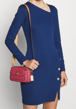 Eng anliegendes blaues Kleid mit langen Ärmeln, goldenen Knopfakzenten und einem hohen Schlitz am Oberschenkel. Rosa Umhängetasche mit strukturierter Oberfläche und goldfarbenen Beschlägen.