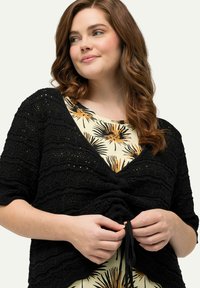 Schwarze gestrickte Strickjacke mit kurzen Ärmeln, strukturiertem Design und Frontbindedetail, getragen über einem hellen Blusenshirt mit floralem Muster.