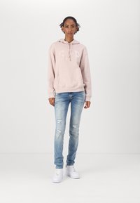Jasnoróżowa bluza z kapturem z logo "GAP", kieszenią, podarte niebieskie dżinsy i białe sneakersy. Prosty design i luźny krój przez całość.