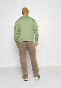 Sweatshirt vert à coupe décontractée, avec poignets et ourlet côtelés, associé à un pantalon en velours côtelé marron et des baskets blanches. Fond uni.