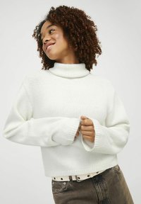 Pull en tricot blanc avec un col montant et des manches longues, doté de poignets côtelés et d'une coupe cintrée. Porté avec un pantalon marron foncé et une ceinture blanche.