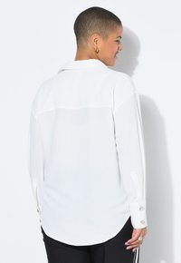 Una persona con el cabello corto lleva una camisa blanca, de espaldas frente a un fondo claro, mostrando los detalles y la silueta de la camisa.