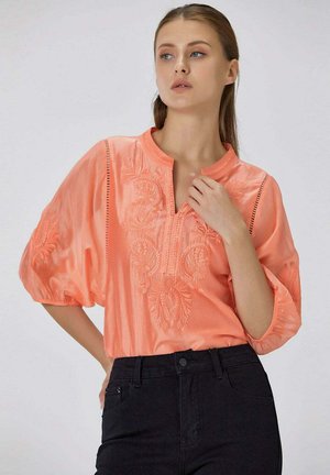 Femme portant un chemisier corail brodé avec des manches bouffantes et un pantalon noir taille haute, posant avec une main près du cou.