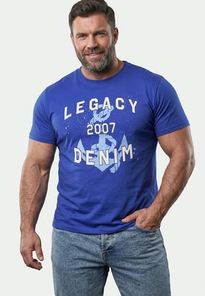 Uśmiechnięty umięśniony mężczyzna w niebieskiej koszulce "Legacy Denim 2007" i jasnoniebieskich dżinsach, z jedną ręką w kieszeni, na jednolitym tle.