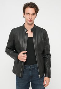 Chaqueta de motorista de cuero negro con cuello alzado, cierre de cremallera y detalles acolchados en los hombros. Lleva puesta sobre una camiseta sin mangas negra.