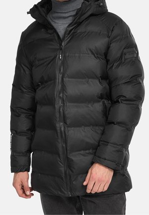 Mann trägt eine schwarze Steppjacke mit hohem Kragen und Frontreißverschluss über einem grauen Rollkragenpullover.