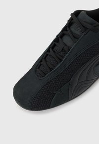 Μαύρο αθλητικό παπούτσι Puma με ύφασμα από δίχτυ, επενδύσεις από σουέτ και σύστημα κλεισίματος με κορδόνια, κοντινή λήψη υπό γωνία σε ουδέτερο φόντο.