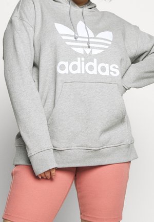 Personne portant un sweat à capuche gris Adidas avec une poche avant et un short de cycliste rose côtelé, debout avec une main sur la hanche.