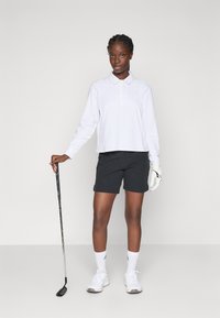 Vrouw in een witte lange mouw polo, zwarte shorts en witte golfhandschoen, die een golfclub vasthoudt, staande tegen een effen achtergrond.