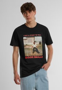 Juodas medvilninis marškinėliai su priekinio vaizdo grafika, kurioje vaikas sėdi ant žolės, su raudonu tekstu "TURIU PASAKĄ" ir "BIGGIE SMALLS."