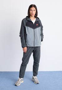 Nike-Jacke in Grau und Schwarz mit Reißverschluss und Kapuze. Kombiniert mit passenden Hosen und hellen Sneakers. Strukturierte Materialien sind sichtbar.