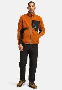 Fleecejacke in Orange mit schwarzen Akzenten, mit durchgehendem Reißverschluss und hohem Kragen. Kombiniert mit schwarzen Hosen und Wanderschuhen mit gelben Details.