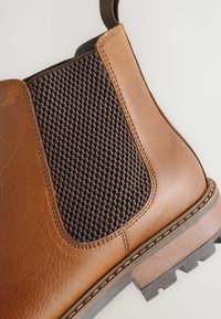 Barna bőr Chelsea csizma sötét textúrájú elasztikus oldalbetéttel és hurok húzófüllel semleges háttér előtt.