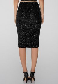 Jupe crayon noire avec un design en sequins texturés. Forme ajustée, longueur genoux, avec une taille mi-haute et une fente au dos. Portée avec des talons noirs.
