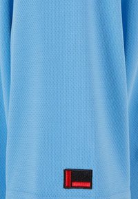 Šviesiai mėlynas tekstūruotas audinys su mažomis perforacijomis. Apatinio kairiojo kampo dalyje yra juodas ir raudonas stačiakampis logotipas.