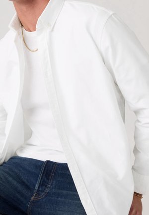 Mand iført en hvid skjorte med knapper over en hvid t-shirt, blå jeans, et guldkædehalskæde og et guldbånd.