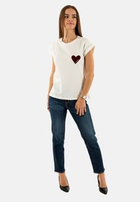 Camiseta blanca de manga corta con un gráfico de corazón burdeos en el lado izquierdo del pecho, combinada con jeans de mezclilla azul y tacones altos negros.